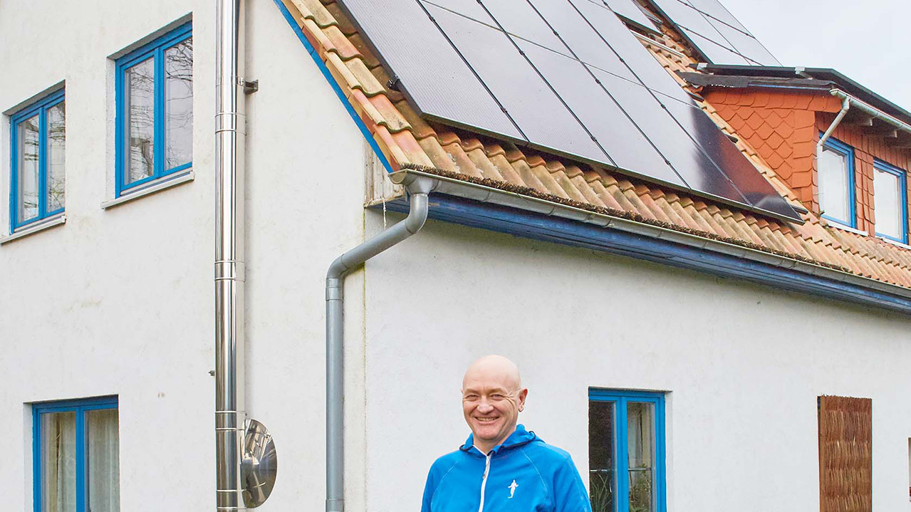 Arthur Meier steht lächelnd vor seinem Haus. Auf dem Dach des Hauses sieht man eine PV-Anlage.